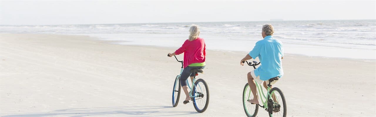 Billedet viser et ældre par, som cykler på stranden.